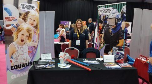 A photo of Giulia Enright smiling to the camera behind a BF/We Are Forging Futures stand at the Summit.